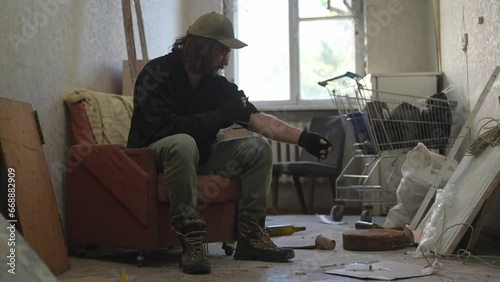 Homeless poor man sitting in a room of an abandoned building filled with his meager belongings. He tightens the tourniquet on his arm and injects an illegal substance, drug into his vein. photo