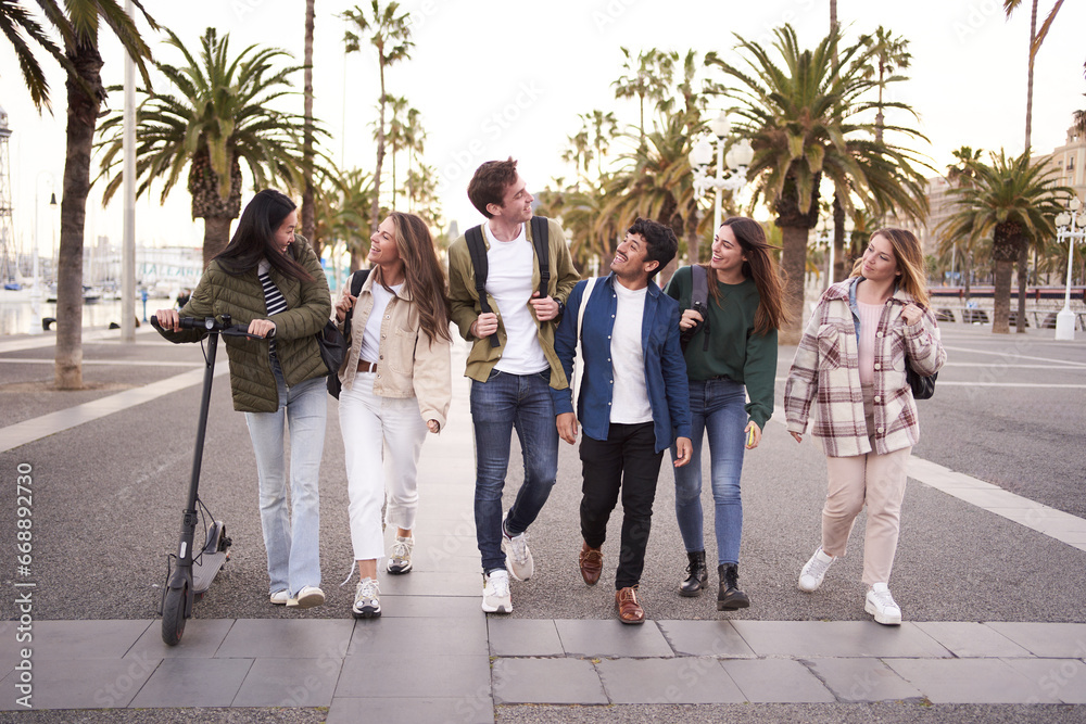 Diverse multicultural group young millennial friends walking along urban street palm trees. University people happy strolling outside on way to campus. Concept of cheerful students together.  - obrazy, fototapety, plakaty 