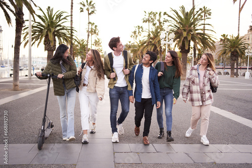 Diverse multicultural group young millennial friends walking along urban street palm trees. University people happy strolling outside on way to campus. Concept of cheerful students together.  photo
