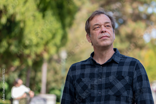 Street portrait of a calm and confident 40-50 years old man wearing a shirt on a neutral blurred background of a city park, space for text, copy space.