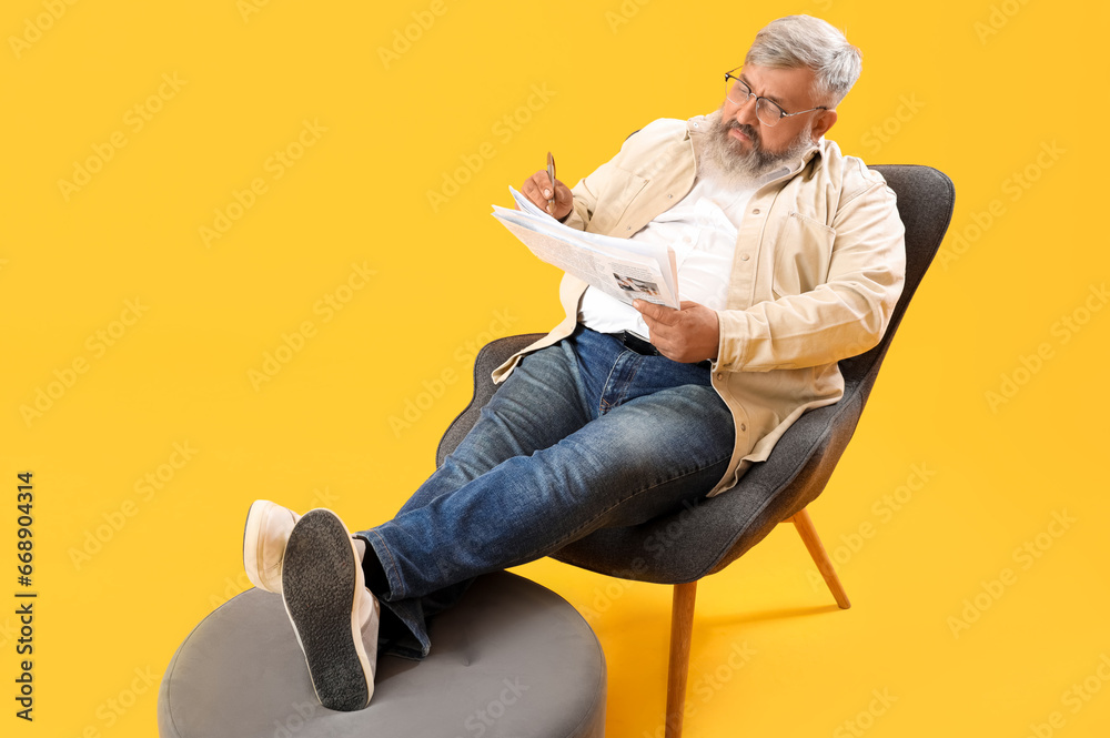 Fototapeta premium Mature man with pen and newspaper sitting on armchair against yellow background