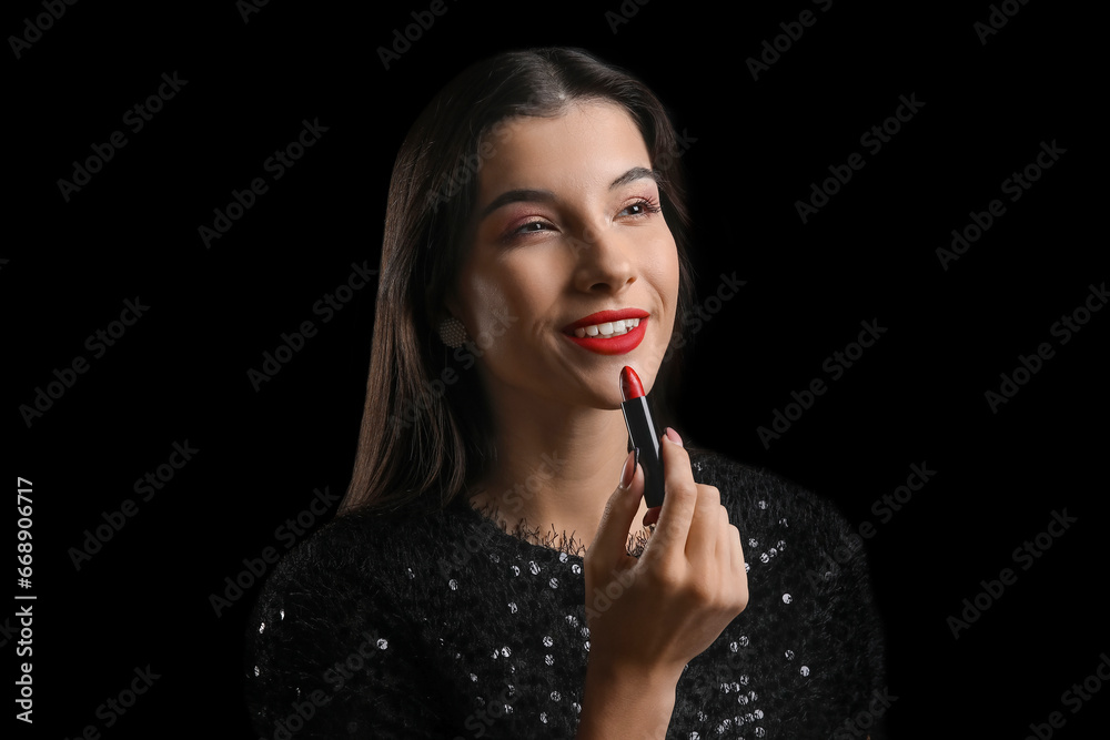 Beautiful young woman applying red lipstick on black background
