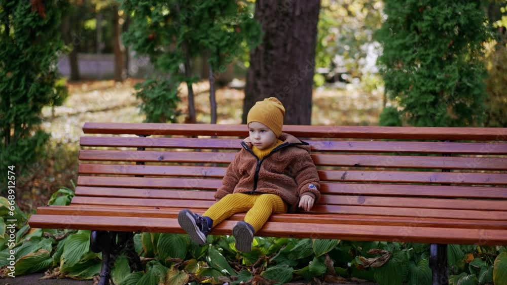 Beautiful toddler boy sits on the bench waving his foot. Calm baby boy resting in the park.