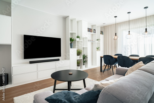 Modern living room with dining area in clean design with builtin furniture and window.There're large TV with sound bar,coffee table,many decorative plants,massive table with chairs and ceiling lights.