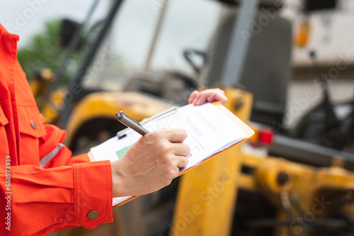 A mechanical engineer is using checklist form to verify the excavator or earthmover machine (as background) condition before operating at construction site. Industrial working scene.