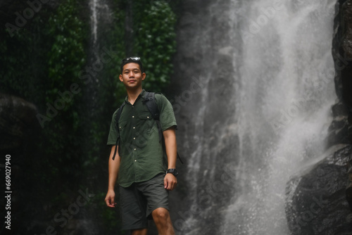 Portrait of man traveller with backpack standing in front of the tropical waterfall surrounded by lush green vegetation