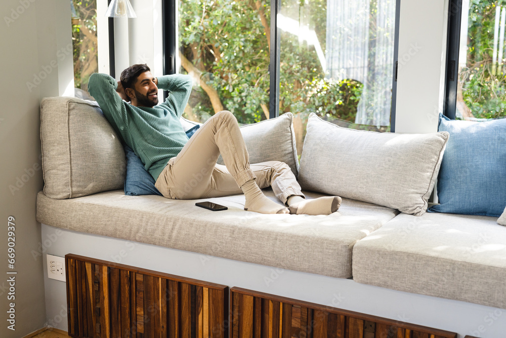 Happy biracial man sitting on sofa with smartphone and smiling in sunny living room Happy biracial man sitting on sofa with smartphone and smiling in sunny living room