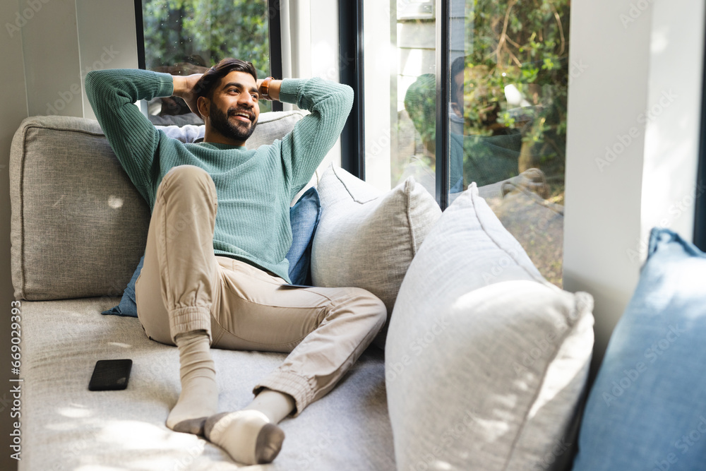 Happy biracial man sitting on sofa with smartphone and smiling in sunny living room Happy biracial man sitting on sofa with smartphone and smiling in sunny living room