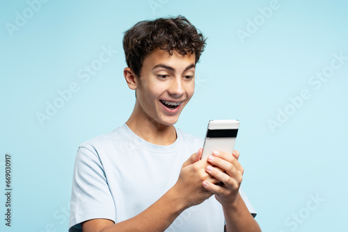 Portrait of overjoyed teenage boy with braces holding smartphone playing mobile game, communication online isolated on blue background. Technology, innovation concept 