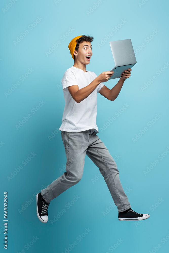 Overjoyed boy teenager holding laptop computer playing video game,  jumping isolated on blue background. Modern guy shopping online with sales. Innovation, technology concept