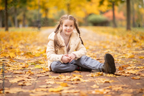 Beautiful girl close-up. A girl sits on autumn leaves in the park in autumn. Portrait of a child outdoors. The girl smiles happily. 
