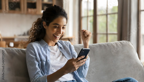 Happy excited young latin woman relaxing on couch using phone winning money in online app  photo