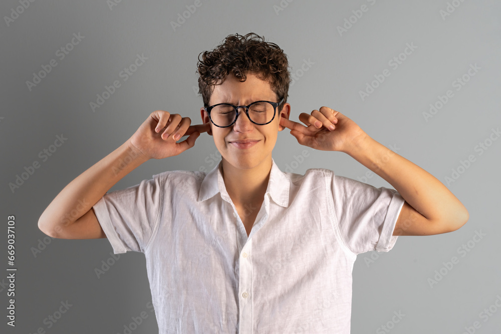 Disturbed by Noise - Curly-haired pre-adolescent boy with glasses squeezing eyes shut and covering ears, displaying a clear sense of discomfort from loud noise or disturbance against a muted backdrop. Disturbed by Noise - Curly-haired pre-adolescent boy with glasses squeezing eyes shut and covering ears, displaying a clear sense of discomfort from loud noise or disturbance against a muted backdrop.
