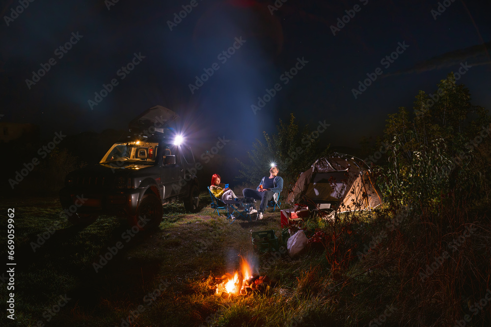 Night starry sky. Smoke and headlamp light. Vehicle near campfire. Couple man and woman sitting near bonfire under majestic blue sky with stars. camping, travelling tourist things, tent chairs, table.