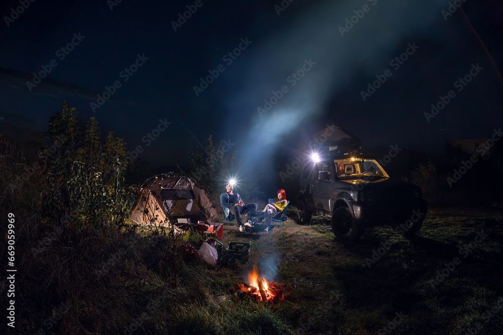 Obraz premium Night starry sky. Smoke and headlamp light. Vehicle near campfire. Couple man and woman sitting near bonfire under majestic blue sky with stars. camping, travelling tourist things, tent chairs, table.