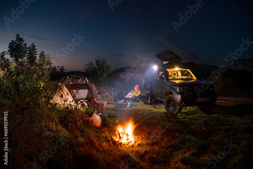 A woman with red hair sits on a folding chair at a table. Uses a smartphone. Camping, bonfire, tent and off-road vehicle. Beautiful sky, sunset.