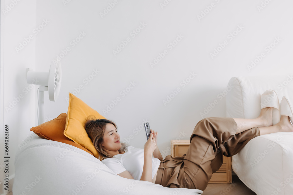 Φωτογραφία Asian Thai woman lying on bean bag sofa, happy chilling spending time alone at apartment room, holding and using phone for texting or video calling with boyfriend, watching series
