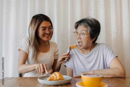 Asian old woman mother and adult daughter smiling, one giving and feeding mom eating croissant bakery, spending time together in cafe, happy good life.