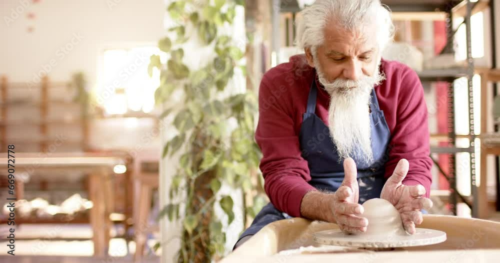 Focused senior biracial potter with long beard using potter's wheel in pottery studio, slow motion