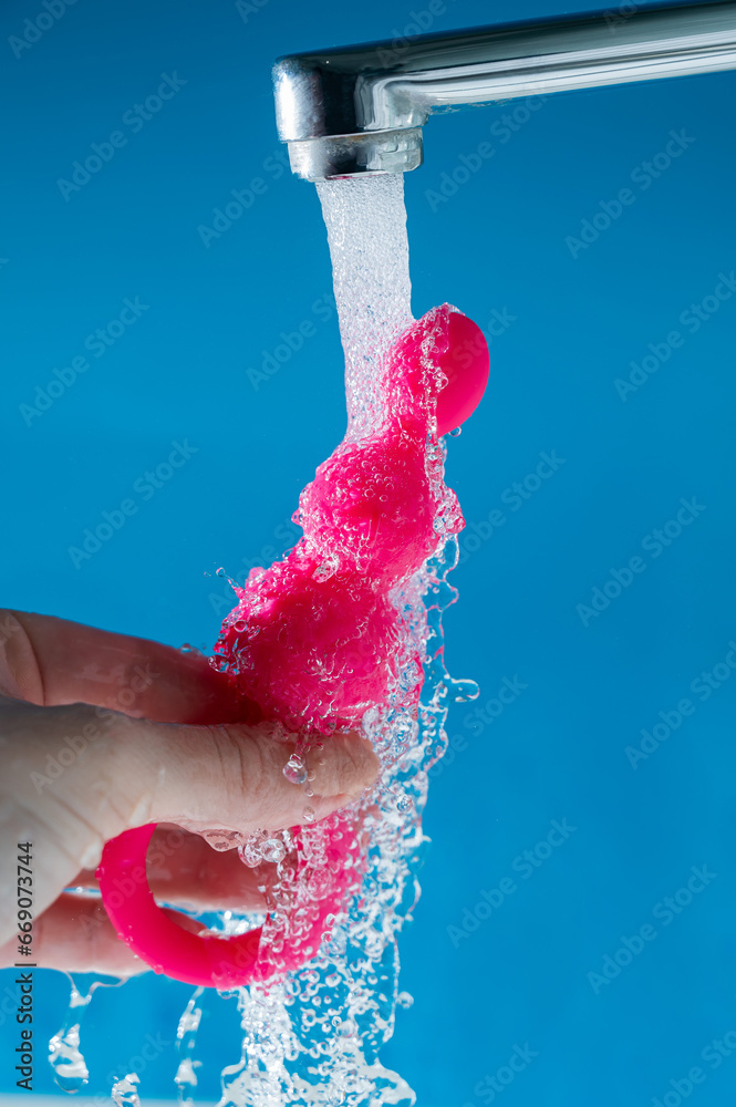 Fototapeta premium Woman holding pink anal beads under running water on blue background. Sex toy hygiene concept.
