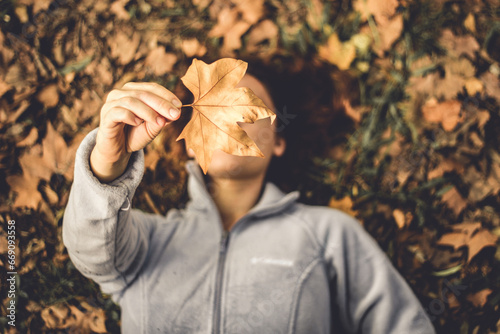 mulher jovem não identificada deitada sobre folhas secas de outono segurando uma folha de plátano em frente ao rosto photo