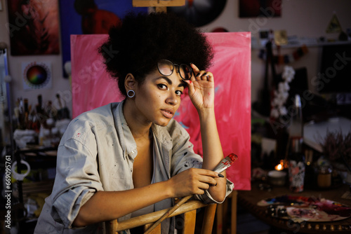 African american female artist holding eyeglasses and paintbrush near blurred canvas in workshop