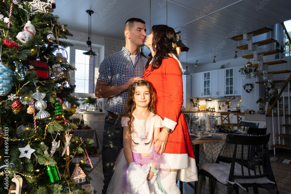 Obraz premium A traditional family of mom, dad and daughter near the Christmas tree in the interior of their house. Celebrating the New year with your family