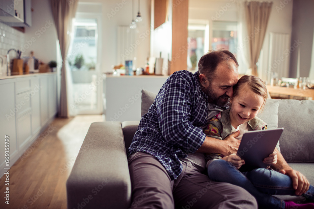 Joyful little girl using the tablet with her father on the couch at home