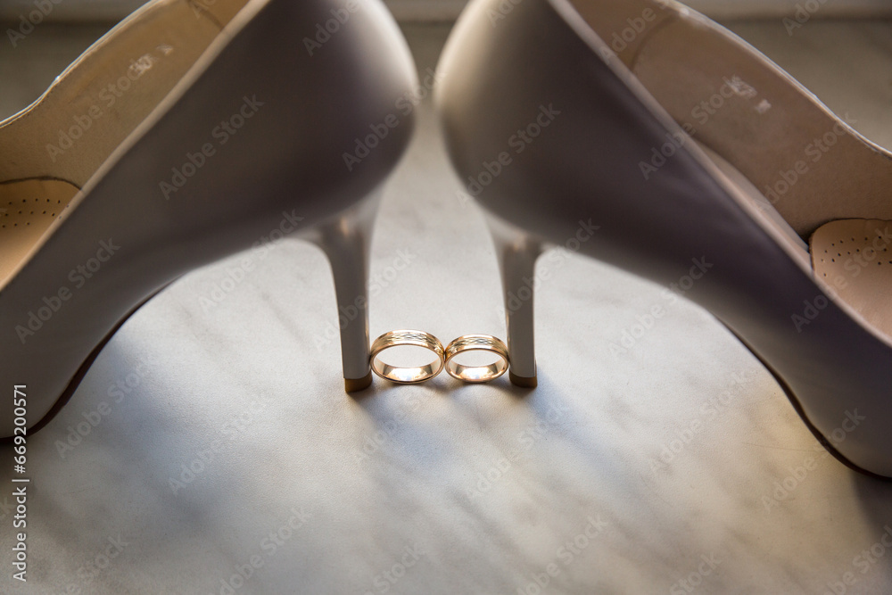 Elegant beige heels beside gold wedding rings, lit by soft natural light. Top-down shot capturing sophistication and romance.