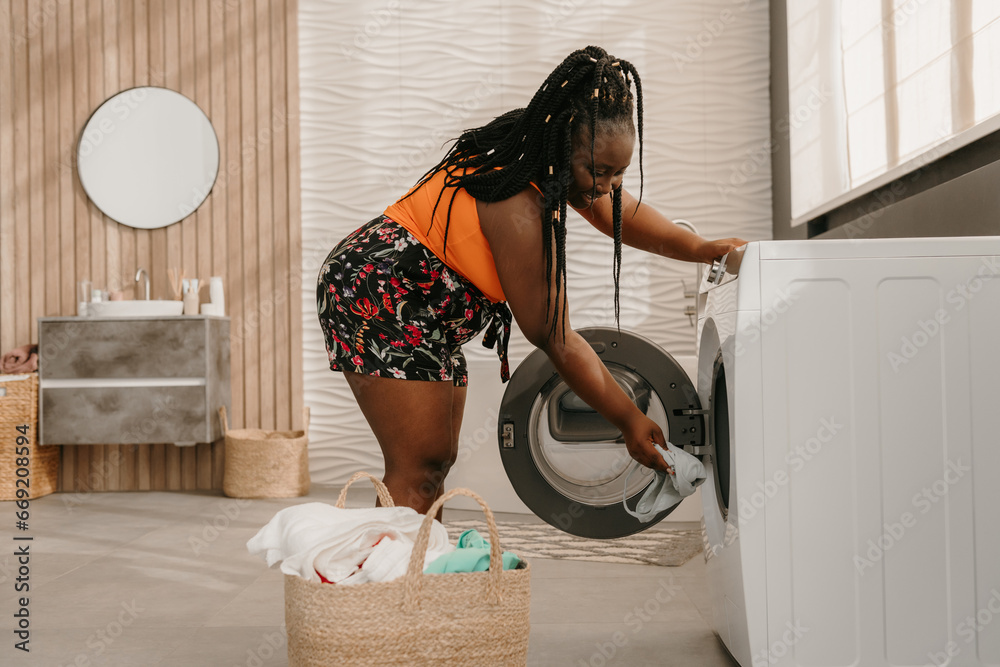 Beautiful curvy African woman loading clothes into washing machine while standing in bathroom - obrazy, fototapety, plakaty 