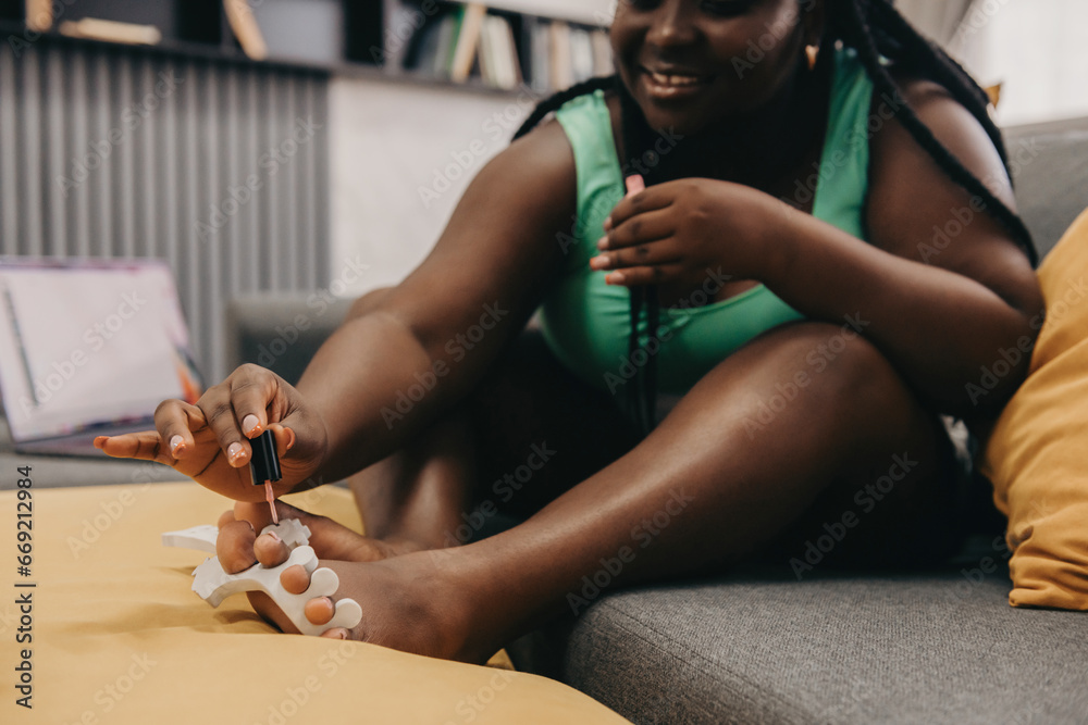 Obraz premium Close-up of joyful plus size African woman painting toenails while sitting on the couch at home