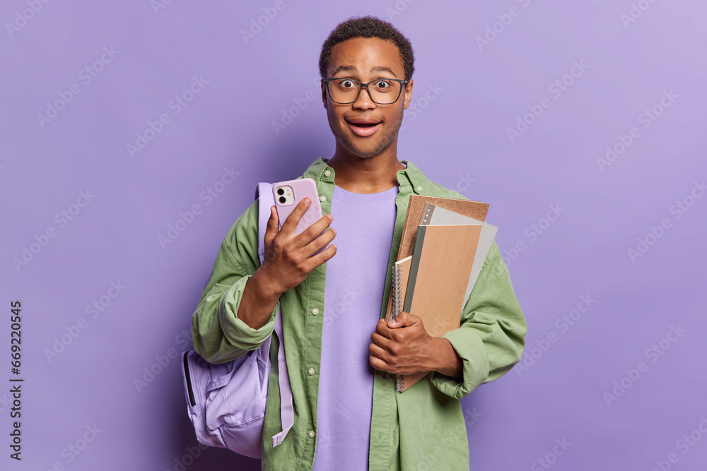 Impressed clever African student with curly hair poses with smartphone and notepads carries rucksack with all necessary things for studying isolated over purple background finds out shocking news Impressed clever African student with curly hair poses with smartphone and notepads carries rucksack with all necessary things for studying isolated over purple background finds out shocking news