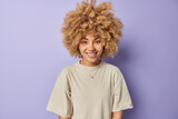 Indoor shot of cheerful curly haired millennial girl smiles happily dressed in casual t shirt looks directly at camera expresses positive emotions isolated over purple background. People and happiness