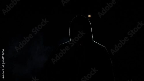 Silhouette of a person on a black background. A man in a hood, illuminated by light, blows. A hacker in the night, a mysterious person whose face is not visible. He smokes a cigarette