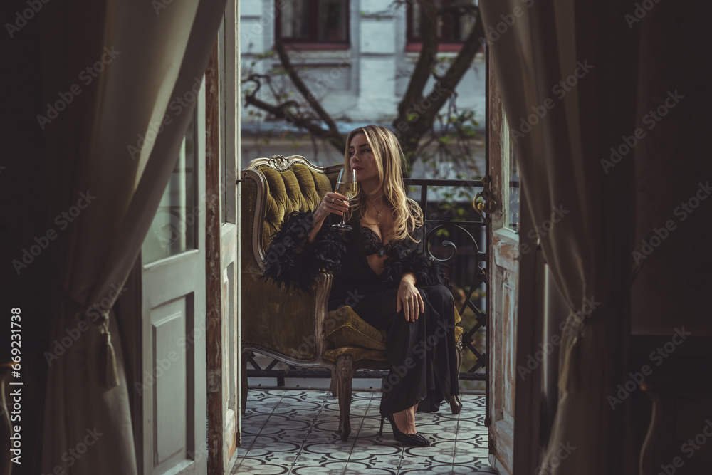 Lady in peignoir with a glass of wine. Sensual photo of European Blonde woman in black silk robe with feathers sleeve. Fashionable concept Lady in peignoir with a glass of wine. Sensual photo of European Blonde woman in black silk robe with feathers sleeve. Fashionable concept