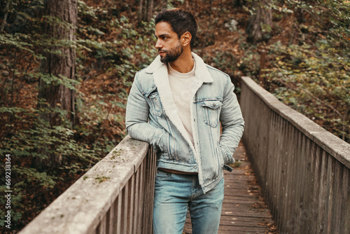 A charismatic young man showcasing style in a denim jacket and jeans, striking a confident pose on an outdoor wooden bridge © rdrgraphe