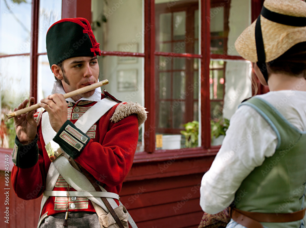 Young redcoat soldier playing flute for a young girl dressed up in old-fashioned clothes.