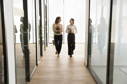 Two multiethnic office employees women walking on hallway, talking. Younger and elder colleagues discussing teamwork, career in successful company, working on business project. Full length shot