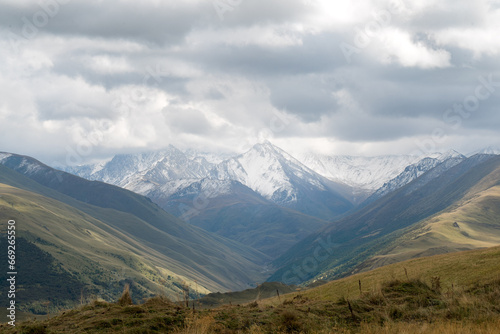 White snowy mountain peaks with low hanging clouds. Green valleys and meadows in the front