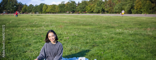 Vertical shot of smiling asian girl sitting on blanket with ukulele guitar, relaxing on sunny day outdoors, resting in park