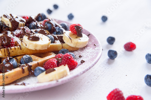 Tasty belgian waffles with blueberries, raspberries, bananas and sugar powder are filled with liquid chocolate.