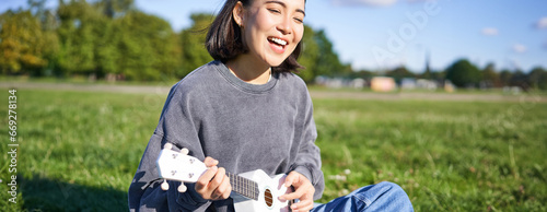 Portrait of beautiful asian woman singing, playing ukulele guitar in park, sitting alone on grass on sunny day photo