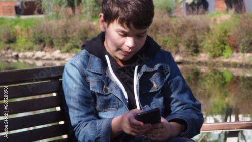 smiling happy fun teenage schoolboy boy sits in a park on a bench use mobile cell phone chat online rest relax outdoors Urban. spending time on social networks, online training, conference. video call