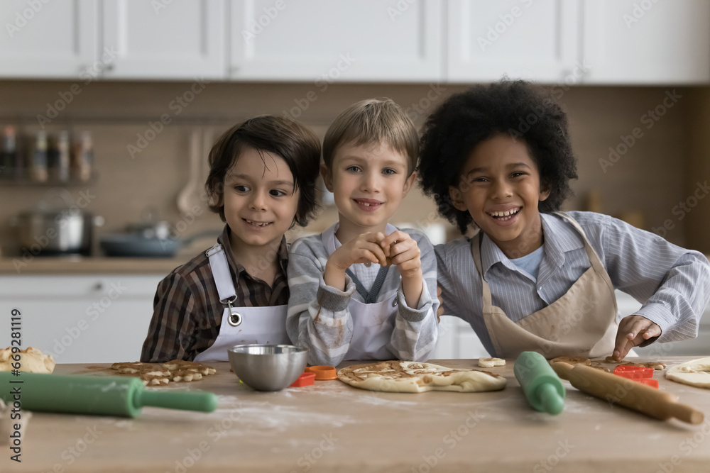 Three multiethnic preschoolers boys in aprons cooking together in kitchen, prepare surprise for family, make dough for homemade pizza posing smile look at camera at home. Cousins friendship and hobby Three multiethnic preschoolers boys in aprons cooking together in kitchen, prepare surprise for family, make dough for homemade pizza posing smile look at camera at home. Cousins friendship and hobby