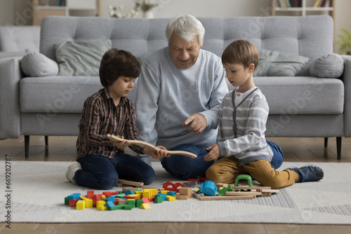 Mature loving great-grandfather spend time with cute little great-grandsons, sit on floor play wooden railroad enjoy playtime at home, having pleasant weekend time and communication. Family pastime