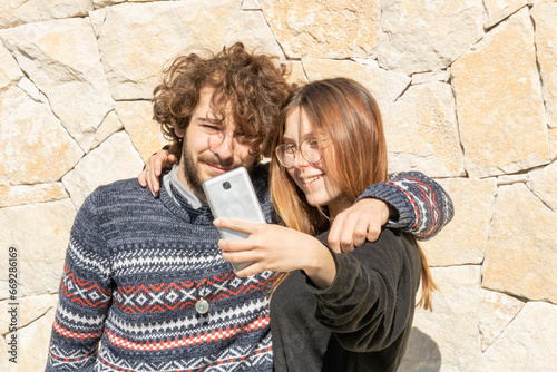 2 jeunes un homme et une femme caucasiens lui brun et frisé elle blonde aux cheveux longs, prenant la pose devant l'objectif photo du smartphone pour un selfie souvenirs entre amis         photo