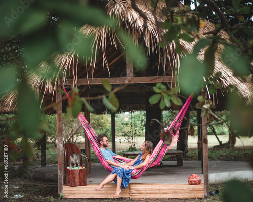 casal jovem deitado em rede cor de rosa em frente a uma maloca com teto de palha rodeado de árvores verdes, em Soure, ilha do Marajó  photo