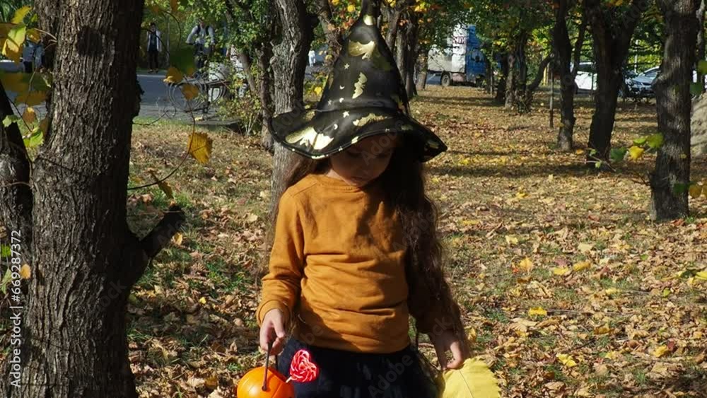 happy laughing child girl in witch costume to halloween. with a jack-o'-lantern candy bowl, trick or treat, in a witch's hat, with colorful balls in the autumn forest, eating spider-shaped candy happy laughing child girl in witch costume to halloween. with a jack-o'-lantern candy bowl, trick or treat, in a witch's hat, with colorful balls in the autumn forest, eating spider-shaped candy