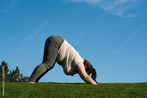 overweight middle-aged woman practices yoga in park, blue sky background. fat woman doing Downward facing dog exercise, adho mukha svanasana pose. Healthy lifestyle, Pilates outdoors in summer photo