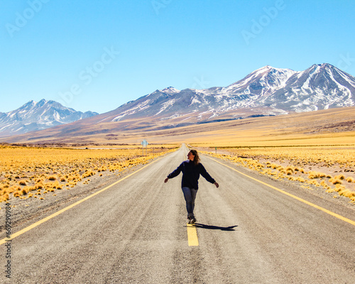mulher caminhando em rodovia no salar do atacama, chile, com montanhas nevadas da cordilheira dos andes ao fundo